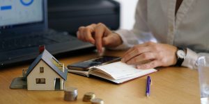 Image symbolique d'une maison miniature posée sur un bureau, entourée de pièces de monnaie, d'une main tenant un stylo, un livre et une calculatrice.